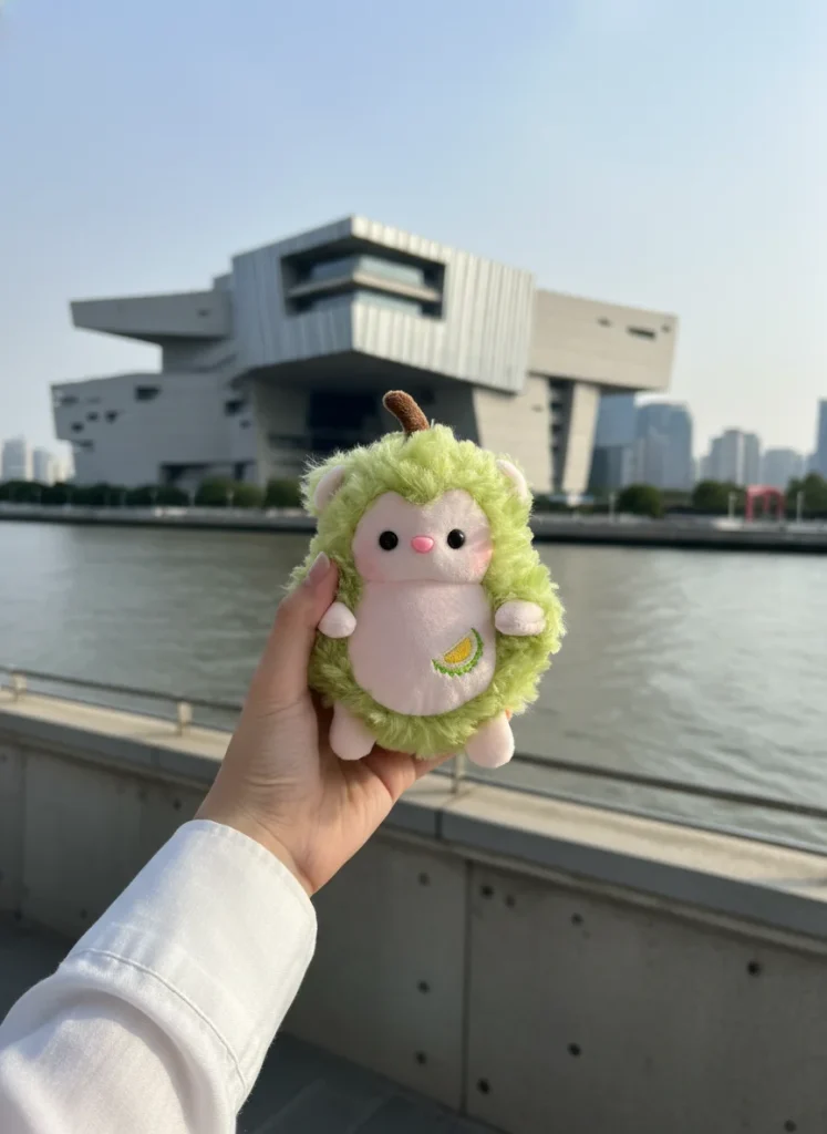 A hand holds a fuzzy green stuffed toy shaped like an animal with a stitched banana on its belly, with a modern architectural building and waterfront view in the background.